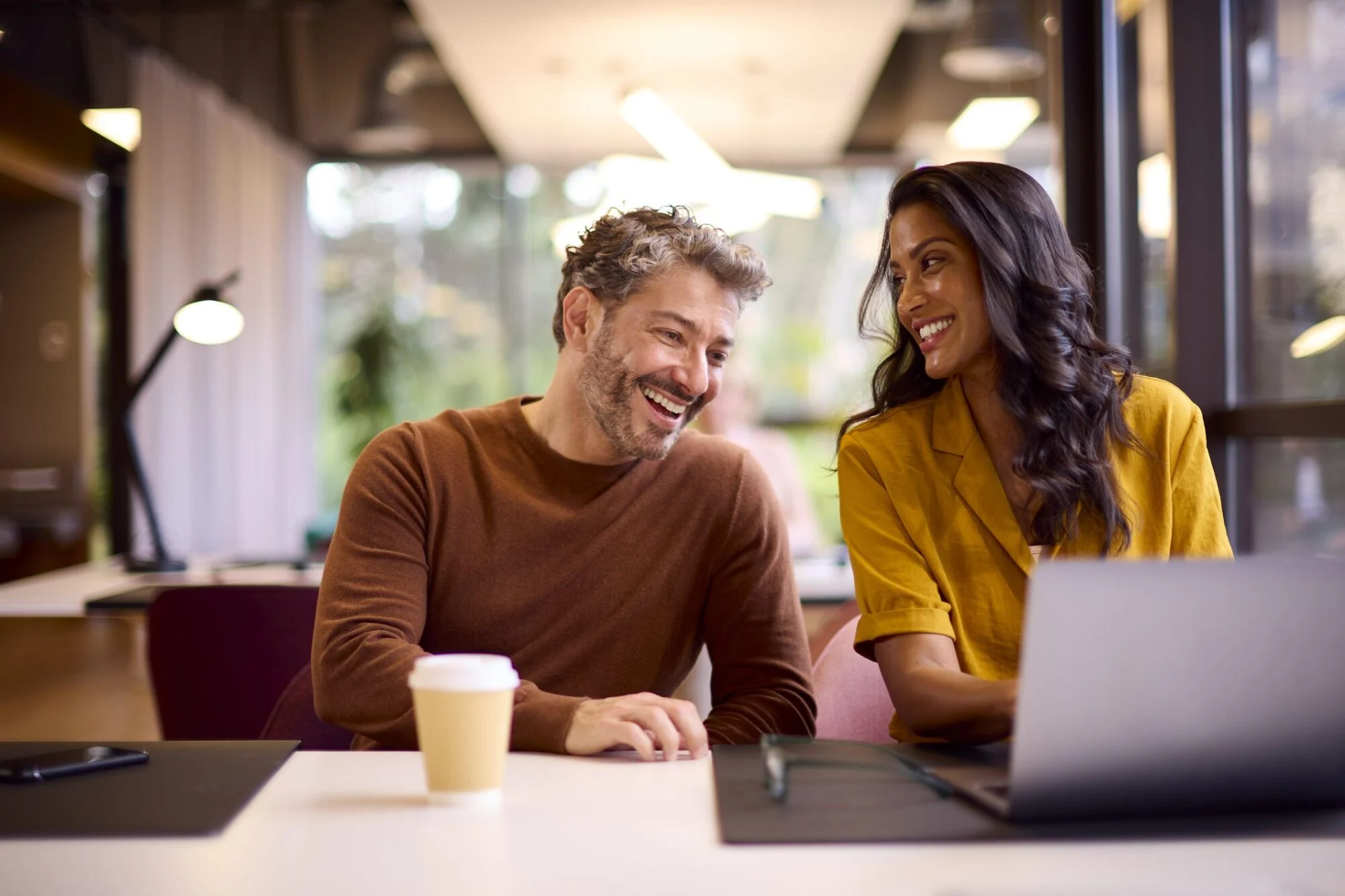 Two colleagues smiling and talking while working together at a desk with a laptop and coffee cup.
