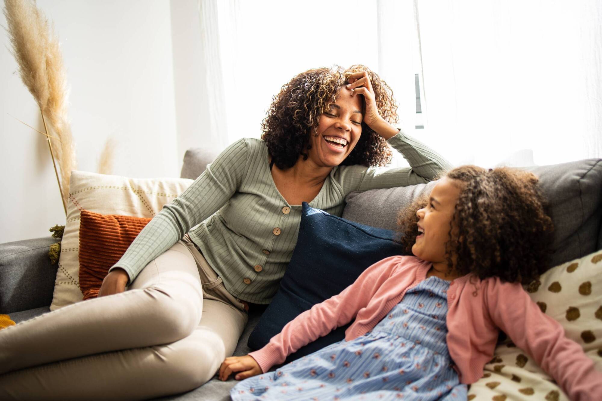 A woman and a young girl sit on a couch, smiling and laughing together.
